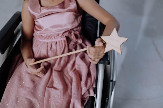 Home A child in a pink dress holding a star-shaped magic wand, seated in a wheelchair.